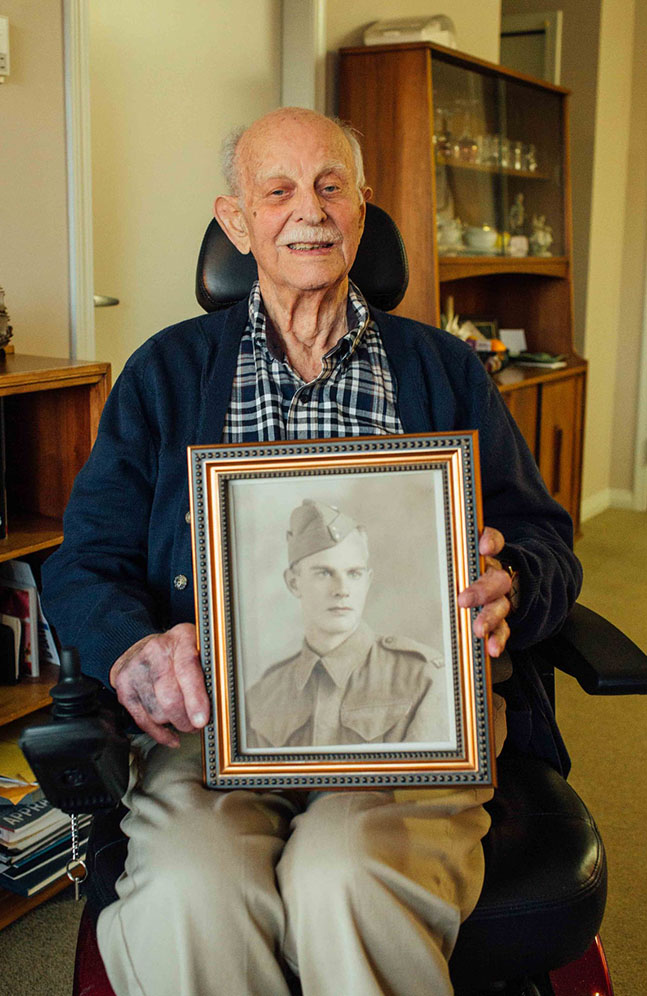 Noel Butcher sitting with photograph of his young self as a WWII soldier